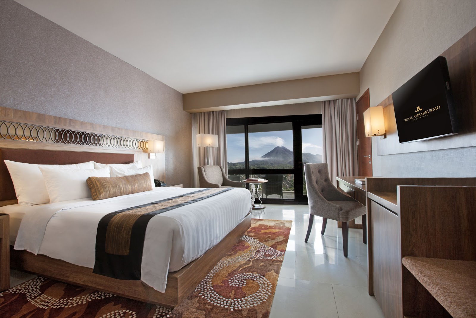 A luxurious guest room featuring a king-sized bed, elegant decor, and a private balcony offering stunning views of a mountain (likely Mount Merapi).