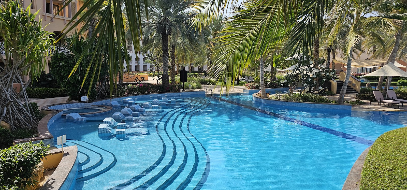 A large, winding swimming pool with various depths and integrated submerged lounge chairs, surrounded by dense palm trees and lush tropical foliage, giving a resort feel.