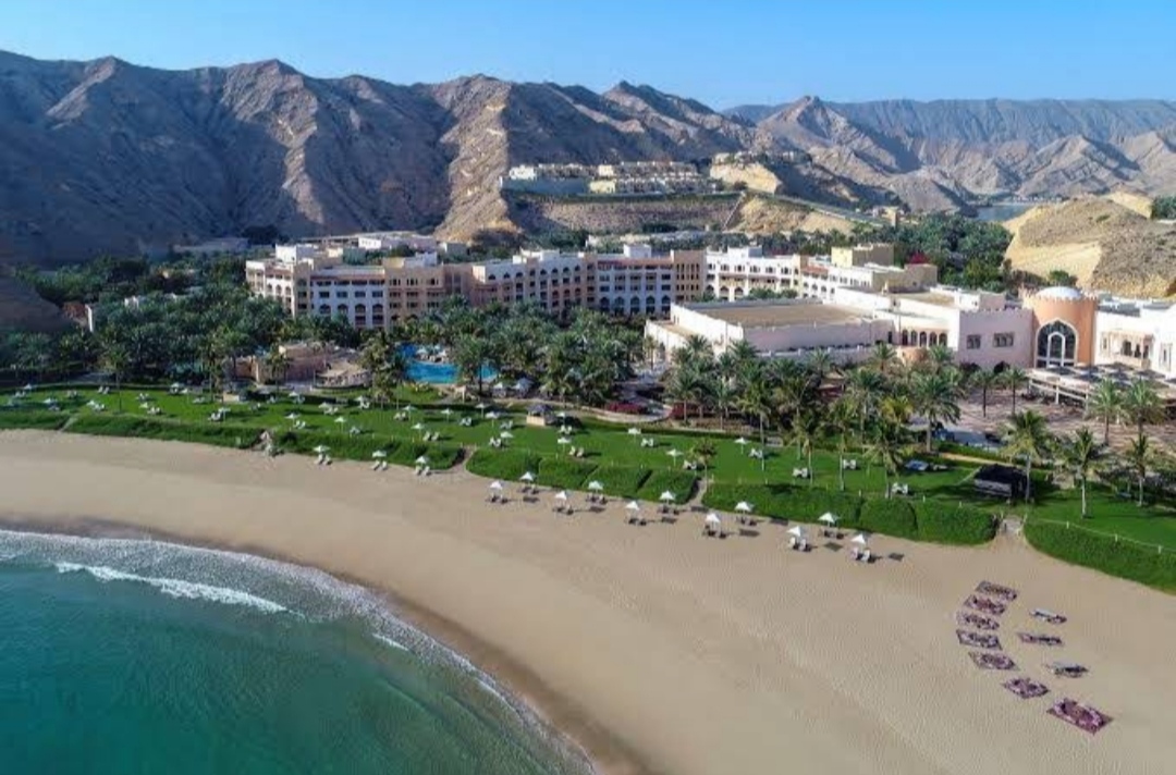An expansive aerial view of the Shangri-La Barr Al Jissah resort, showcasing its multiple hotel buildings, vast green lawns, swimming pools, and long private beach against a backdrop of majestic mountains.