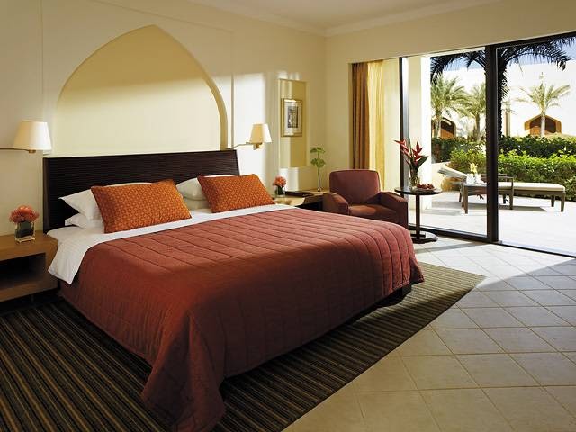 A spacious guest room featuring a king-sized bed, traditional Omani architectural accents like an arched alcove, and direct access to a private ground-floor terrace with lush garden views.