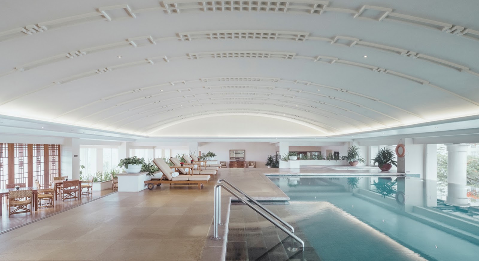 A spacious and elegant indoor swimming pool with a high vaulted ceiling, offering poolside loungers and ample space for relaxation.