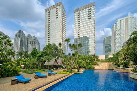 An expansive outdoor swimming pool area surrounded by lush tropical landscaping, sun loungers, and towering city skyscrapers in the background.