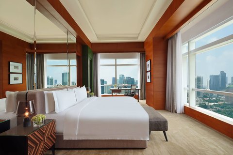 A luxurious hotel bedroom with a large king-sized bed, elegant wooden wall finishes, and expansive floor-to-ceiling windows offering panoramic city views, including a desk area.