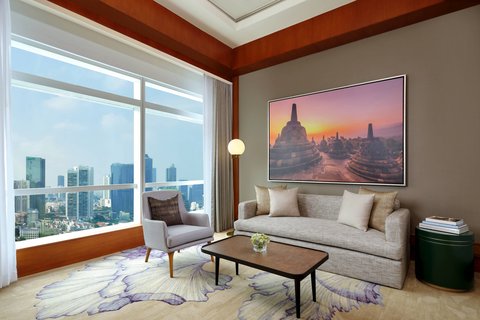 An elegant living area within a hotel suite, furnished with a contemporary sofa, armchair, coffee table, and large windows revealing a city view. A prominent artwork depicting Borobudur temples adorns the wall.